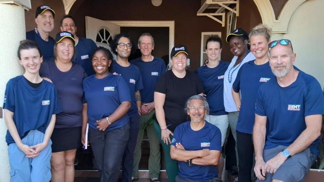 The UK-Med team of a dozen women and men wearing their blue uniform shirts. They smile for the picture on the porch outside their base in Jamaica.