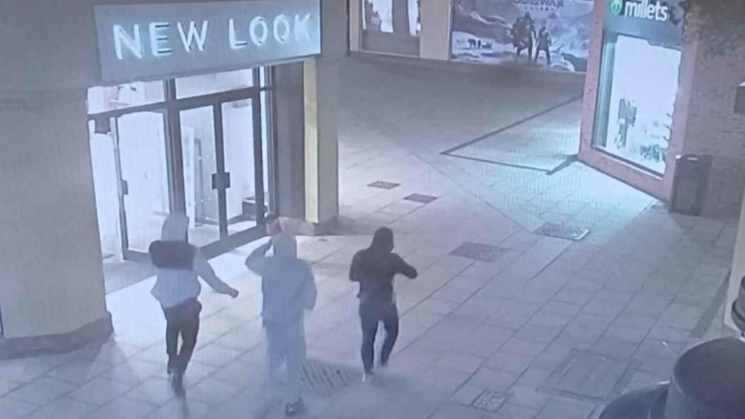 A still from CCTV showing three people in hoodies walking away from the camera with shops either side of them. One is in a white and black tracksuit, another is in all grey and another is in all black.