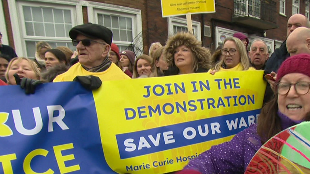 Protesters hold up a blue and yellow banner calling for the Marie Curie hospice to be saved. One of the demonstrators is banging a drum.