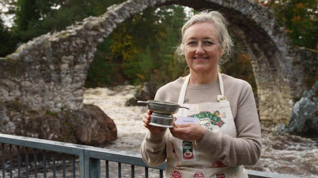 A woman standing outdoors in front of a stone arch bridge, holding a silver cup trophy. she is wearing glasses, a light grey top, and an apron.