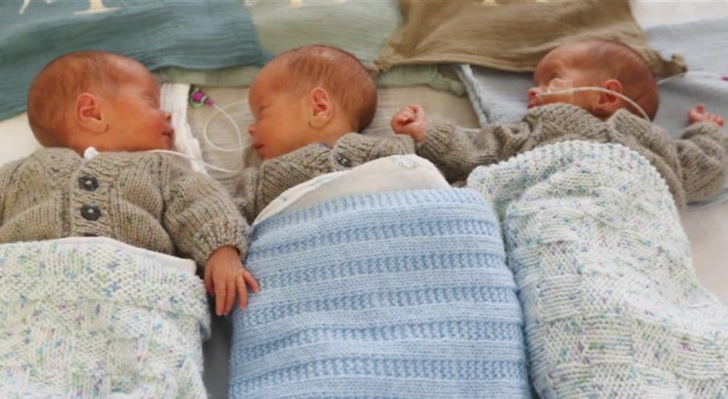 Three newborn babies lie next to each other wrapped in blankets. They are wearing matching jumpers.