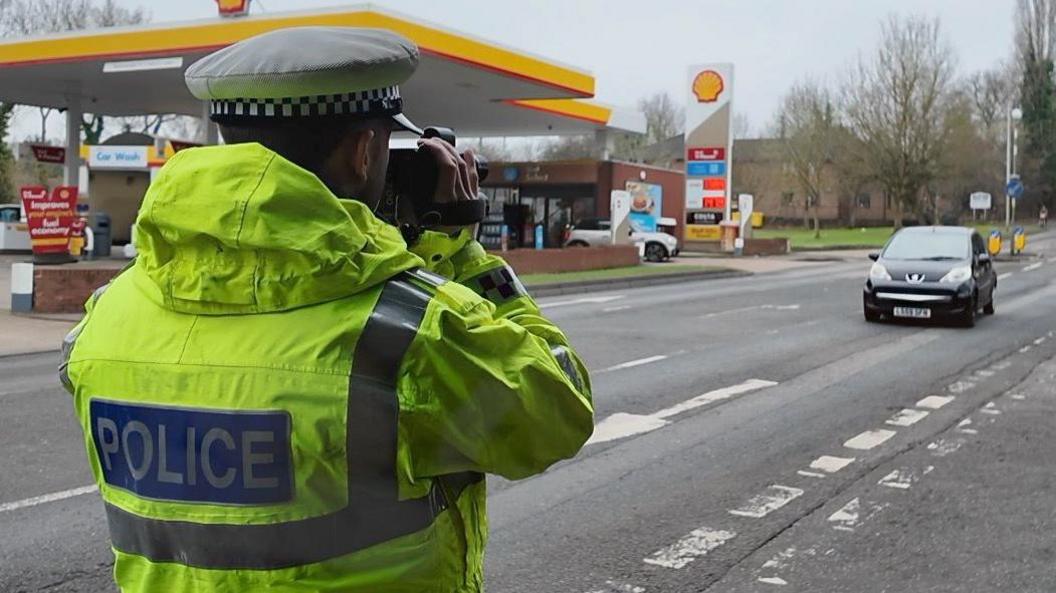 A police officer standing in a yellow high visibility police jacket with a white and black police hat on. He is holding a camera at the side of the road, which can help to detect whether a car is speeding. A car is travelling along the road in front of the officer.