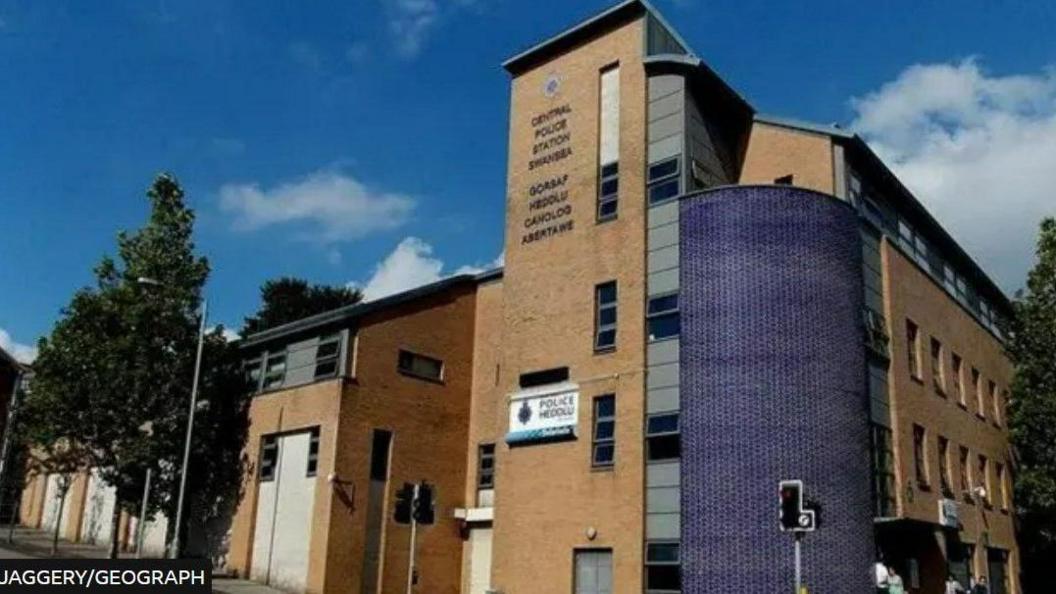 Swansea Central Police Station main entrance seen from the street. The building is light brown brick with a blue rounded facade on the corner and a large police/heddlu sign