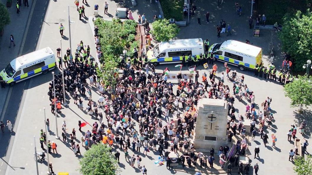 An aerial view of a large crowd of people gathering close to a war memorial. They are partially surrounded by three police vans and groups of police officers. The spaces is dotted with trees.