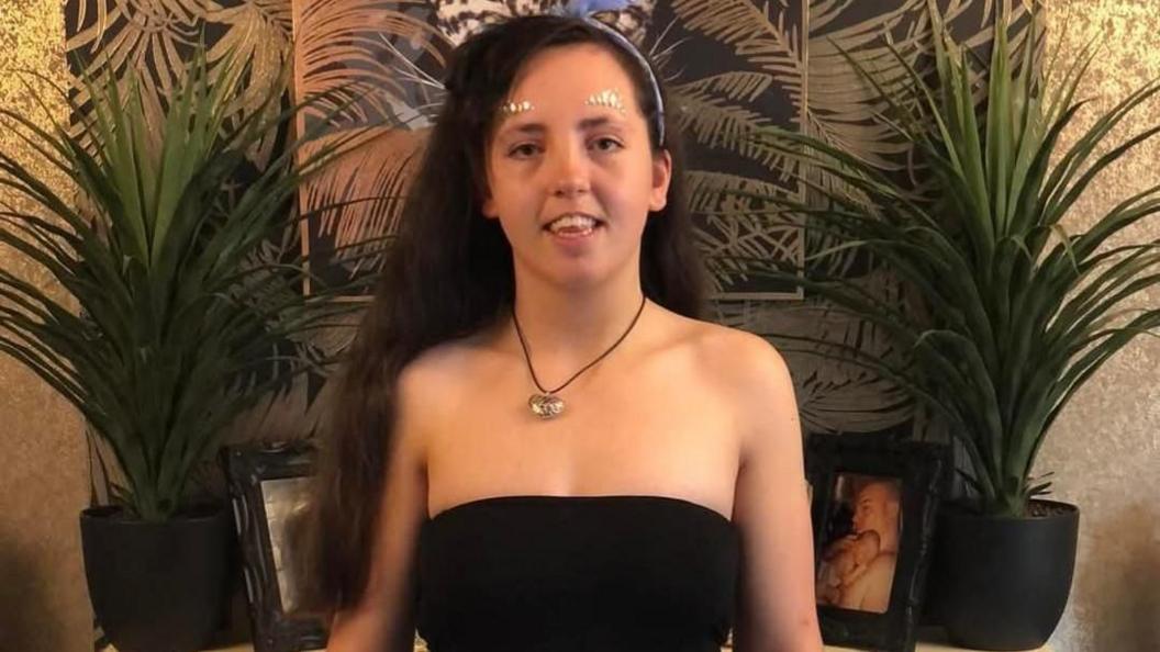 A girl with long dark hair with a headband and wearing a necklace and a black top. She is standing in front of a wall and a cabinet with two plants and pictures on it.