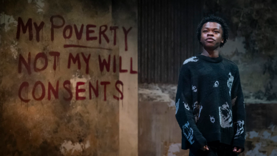 Kyle Ndukuba dressed in a black jumper in stood in a dilapidated warehouse with the words 'my poverty not my will consents' spray painted on the wall