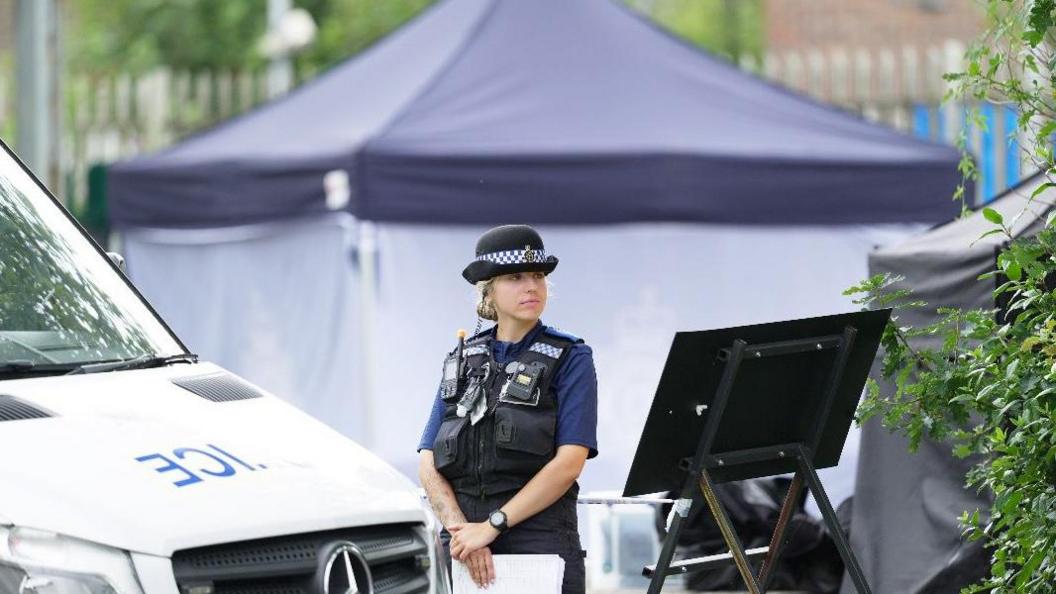 A blonde, female police officer in her uniform standing between a police van and a sign. In the background you can see a navy police tent.