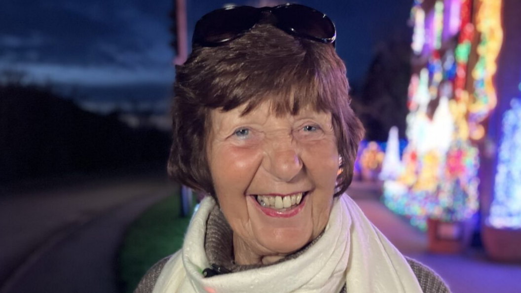 Margaret Ryles is smiling at the camera. She has short dark hair and is wearing a pair of dark glasses balanced on her head. She is wearing a cream scarf and a brown jumper. She is stood in front of the lights at night-time.