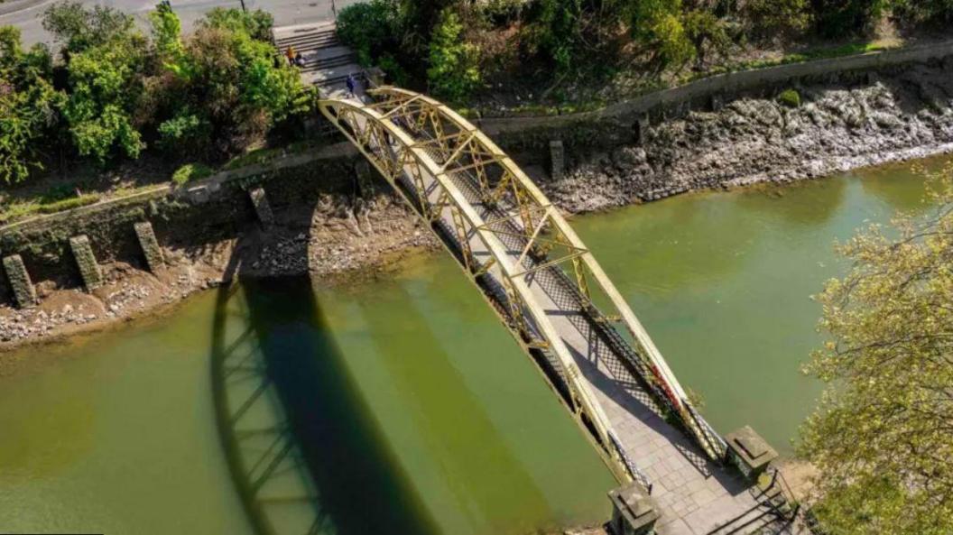 An aerial image of Langton Street Bridge before the repair work started. It is a yellow banana shaped metal bridge. The riverbanks are stripped back to mud and stones and appear somewhat unstable against rising tides.