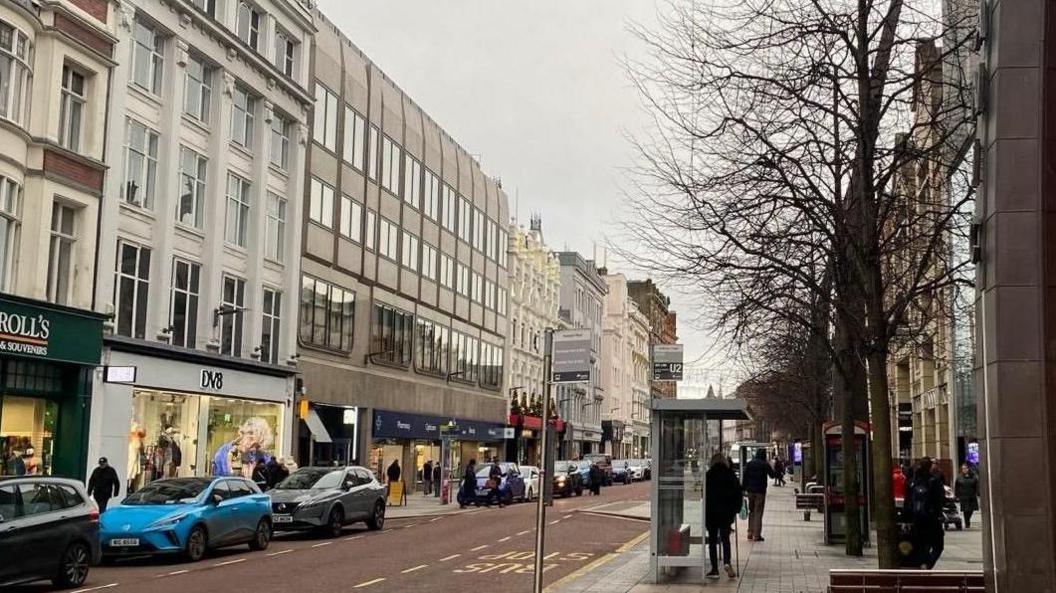 Image of the upper end of Royal Avenue/Donegall Square, there are various people on the pavement on both sides and blue and grey cards to the left.