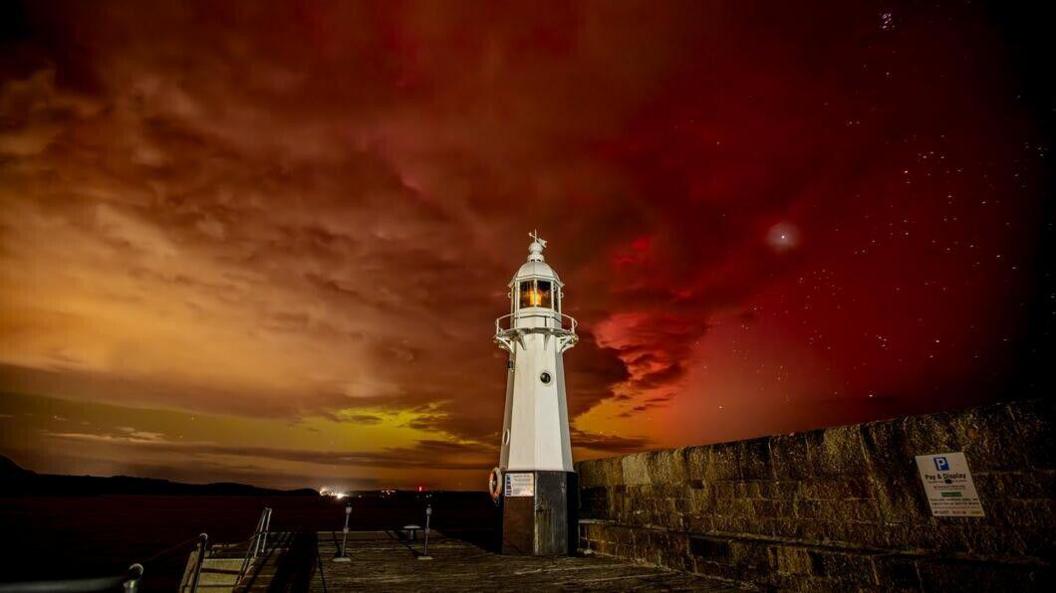 Mevagissey lighthouse is pictured in the front. It is white and surrounded by a brick wall. In the background the sky is orange with hints of yellow within it.