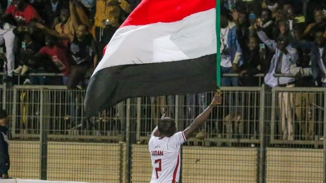 A Sudan player in a white shirt, with his back to the camera, holds up a large red, white and black Sudan flag in front of a stand filled with celebrating supporters