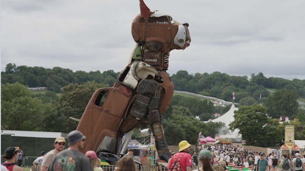 Crowds walk around a statue of a dog made from scrap cars and metal