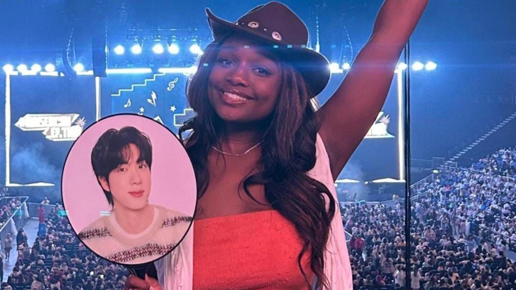Lauryn is wearing a brown cowboy hat, she has brown long wavy hair. She is wearing a red top with a white scarf. She is stood with her back to the stage and concert full of people, holding a face card with BTS member Jin on it. 