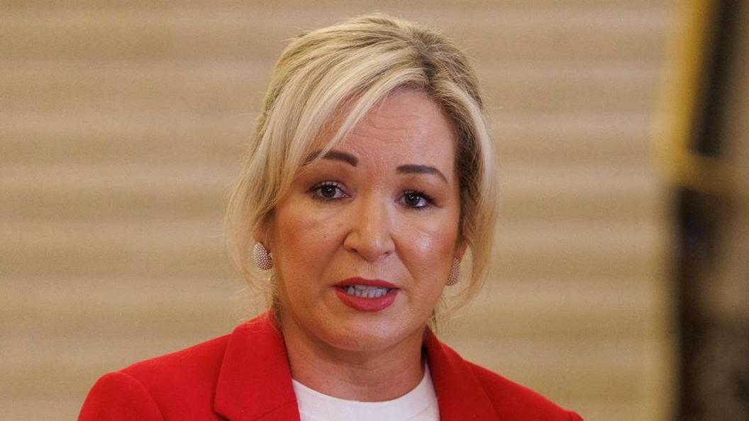 Michelle O'Neill has blond hair tied back and parted to the side and is wearing a red jacket and white t-shirt