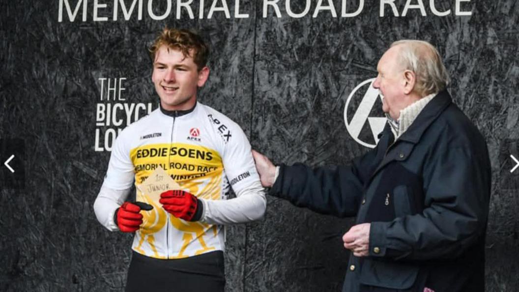 A young man - Aidan Worden - smiles, holding a piece of paper that reads '1st Junior'. He wears a white cycling jersey and red gloves. An man in a black jacket is patting his arm, congratulating him.