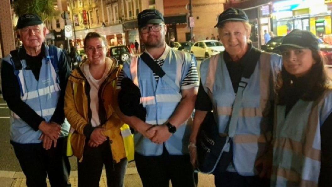 A group of men and women on a night time street. They are weaing blue reflective vests.