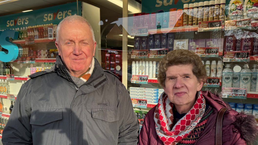 An elderly man with very short white hair wearing a grey jacket standing next to an elderly woman with brown hair wearing a colourful scarf and a purple jacket