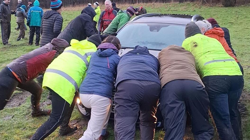 A group of eight people wearing waterproofs pushing a car in a muddy field.