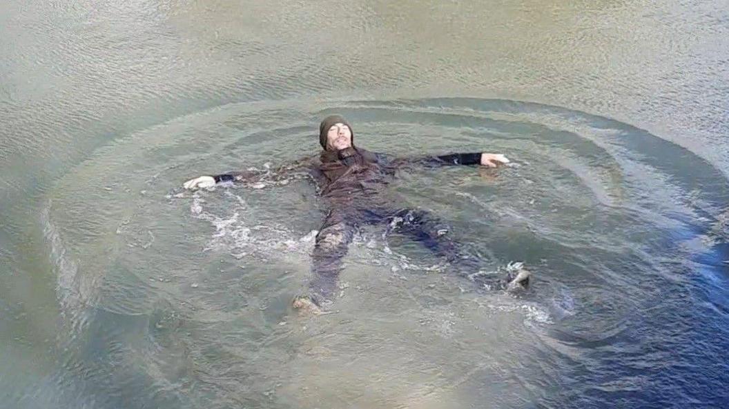 A man in a black jacket and trousers floating in water with his arms and legs spread out. He is wearing shoes and a hat and is demonstrating how to float if you get into difficulty in the water 