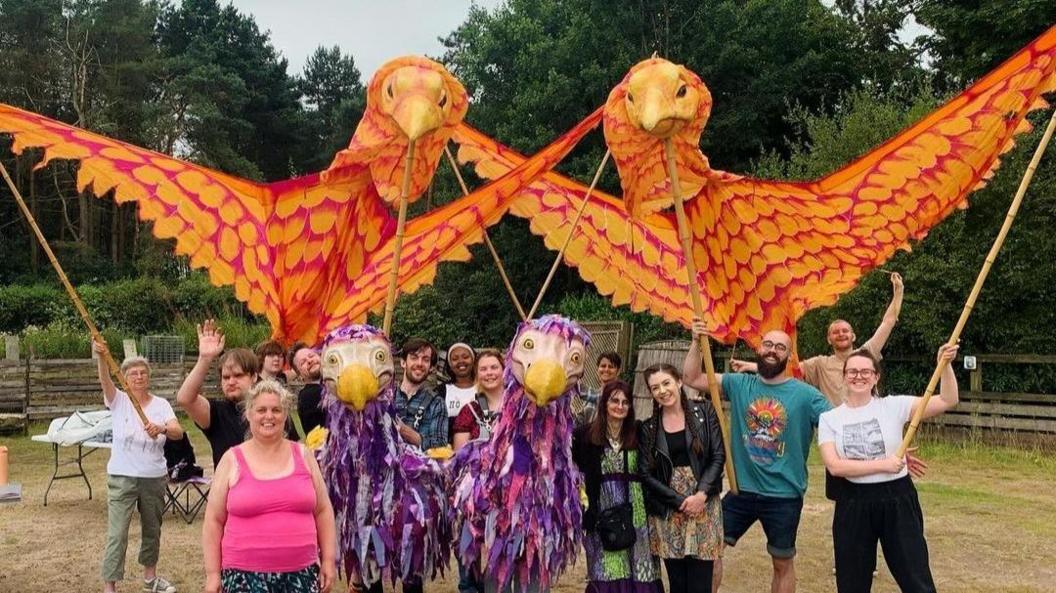 A team of people holding up two large orange bird puppets with wooden sticks, with two of the group dressed in purple emu costumes