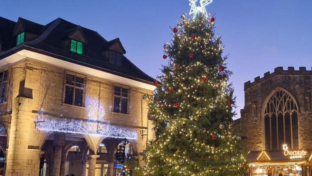 A Christmas tree at dusk in Peterborough city centre. The large tree is covered in lights and has star on top. It stands in front of a two-storey building decorated with twinkly lights.