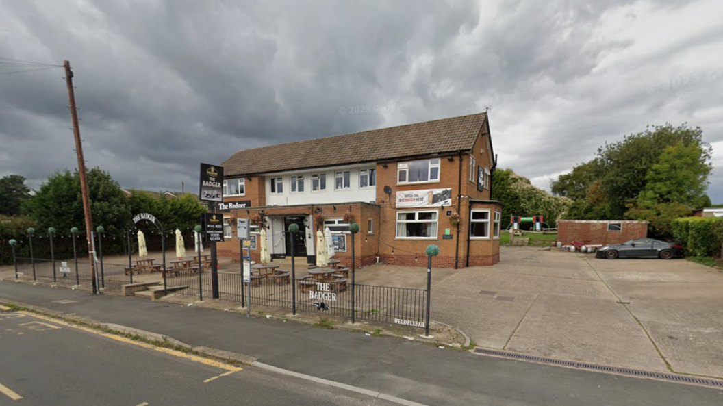 A pub with seating out the front, with grey skies above it. There is an empty car park outside and the road can be seen in the foreground, with a telegraph pole to the left of the shot.