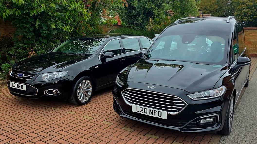 Two black funeral cars are parked on herringbone-patterned block paving, with trees in full leaf behind them.