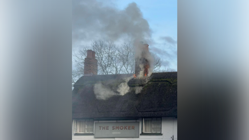 A thatched roof with two chimneys and a pub sign which says the smoker on the front of the white building, between two windows, fire is coming from one of the chimneys