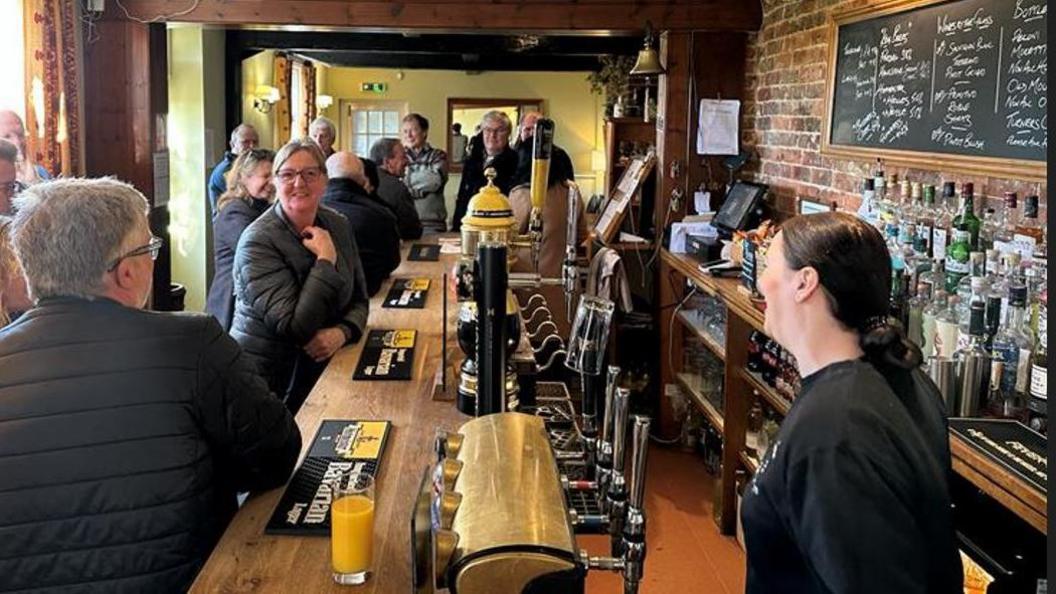 An interior shot of a busy bar area of the Swan on the Green pub in West Peckham with a smiling woman behind the bar serving about half a dozen people sat and propped up the other side of the bar