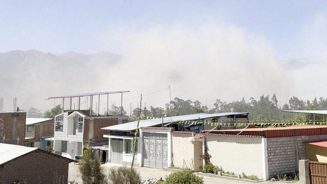 A haze of dust rises from a settlement with one and two-storey buildings. There are mountains in the background.