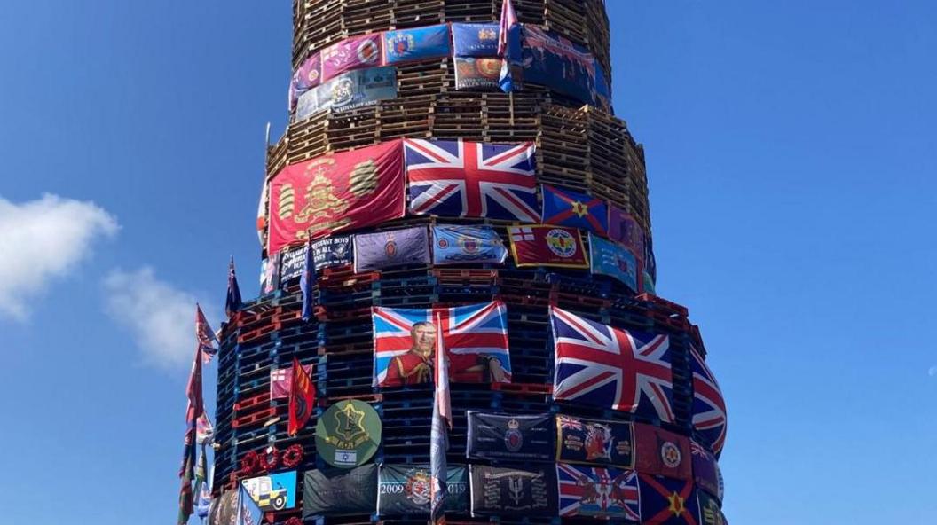 A large number of flags including union flags and a flag depicting an image of King Charles placed on a bonfire in Derry's Bogside.
