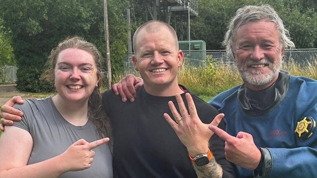 Ant Crocker smiles as he holds up his left hand, which has a wedding ring on the fourth finger. A woman is on the left of the picture, and a man in scuba-diving gear is on the right. Both are pointing at the wedding ring and smiling.