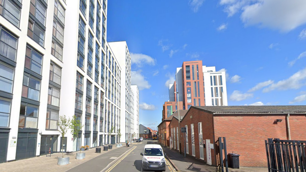 A backstreet in a city centre with high rise buildings and newly planted trees  and a one storey building closer to the camera on one side