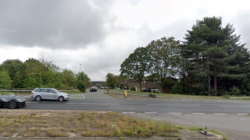 Road junction off a major dual carriageway road. There is a crossing point to the right of the photo where eastbound traffic can cross in front of westbound traffic to enter into the Standens Barns area. Two cars are visible to the left on the main road and one has just turned into the side road. There are trees on either side of the road.