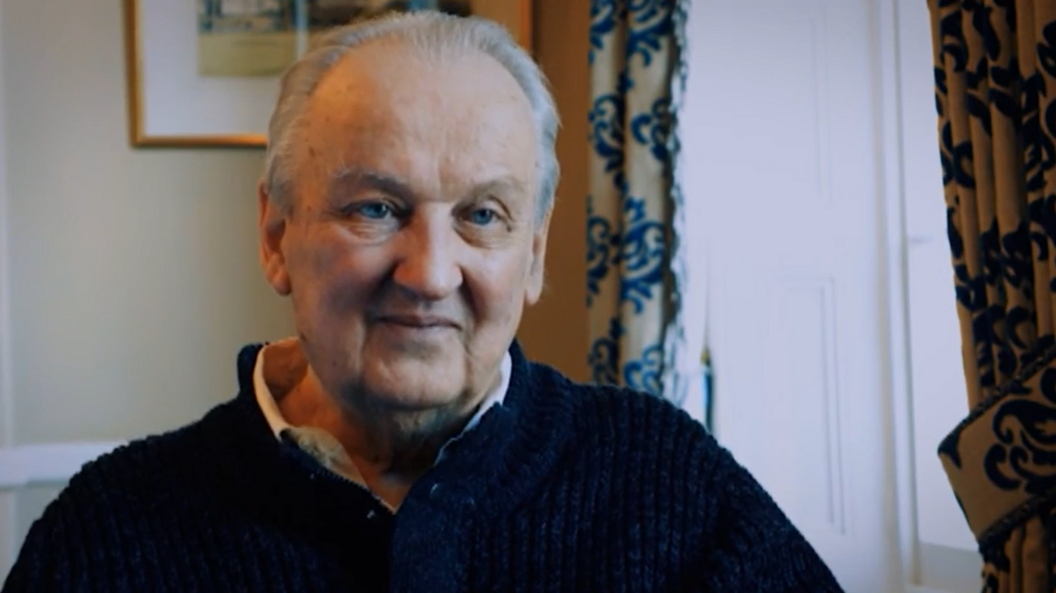 Douglas Gowan wearing a dark knitted jumper sits indoors beside a bright window. Patterned curtains frame the window, while a framed picture hangs on the wall behind. The photograph is warmly lit and focuses closely on his face and upper body.