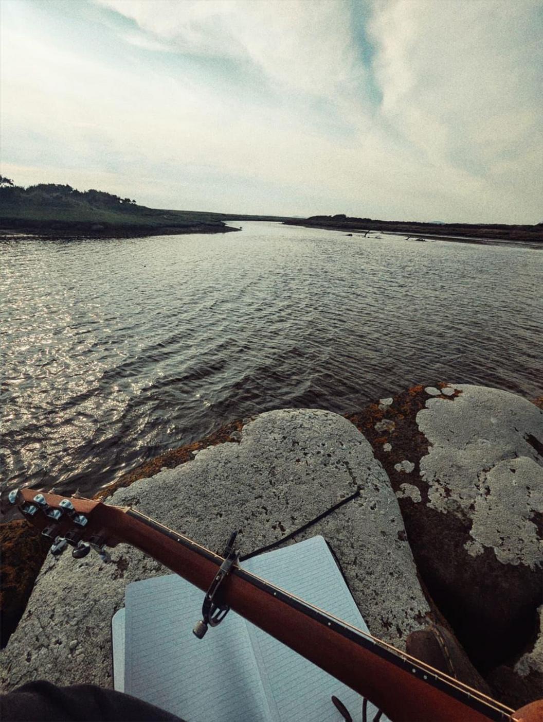 Llun wedi ei dynnu gan gerddor yn eistedd wrth afon yn cyfansoddi ar y gitâr