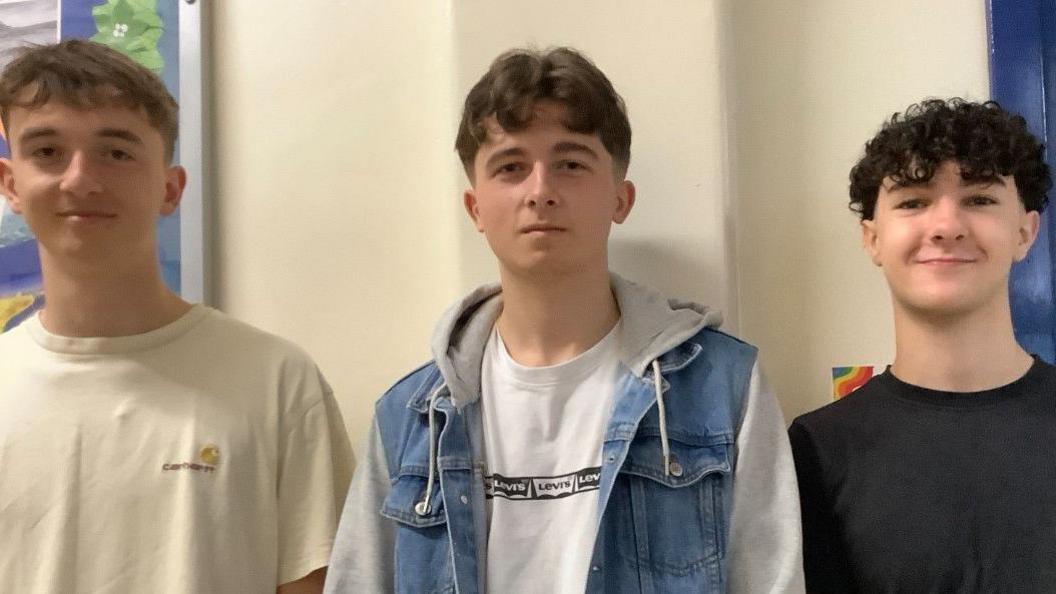 Three young white men, all aged 18, stand side by side in a mixture of coloured t-shirts looking into the camera with slight smiles after recieving their A-level results