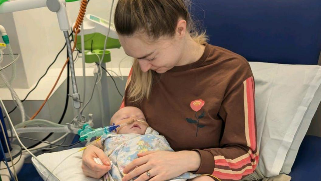 Lauren Auckland sits in a hospital holding her baby Riley, who is on a ventilator.