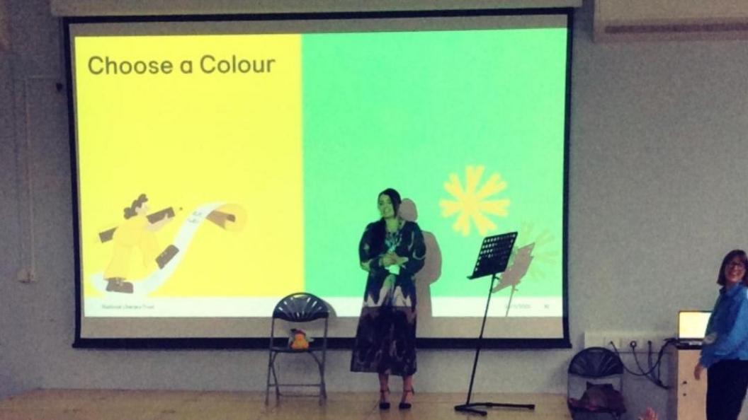 A woman stands in front of a projector screen which is split into two sides. The left has text saying choose a colour on a yellow background and the right has a green background