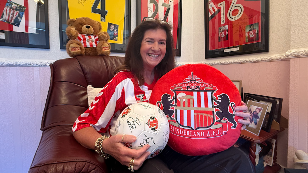 Michelle Barraclough, pictured, has brown hair and is wearing a red and white Sunderland shirt, while holding up a large red Sunderland AFC branded cushion and a white football, which has been signed by various footballers. Behind her are four framed football shirts.