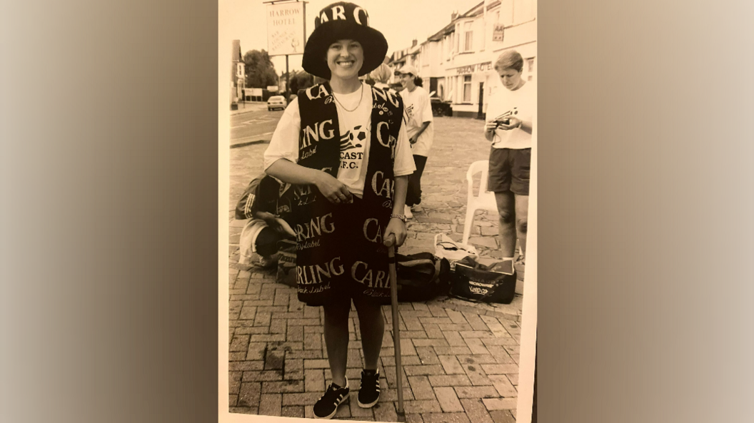 Pauline Fletcher wearing a Carling branded bucket hat and scarf, with a walking stick, standing on a cobbled pavement. 
