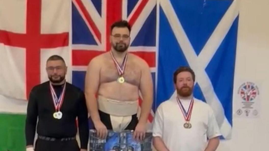Three medal-winning athletes stand on a podium at the British Sumo Championships. The central figure, on the highest tier, represents first place; the person on the left is second, and the one on the right is third. All wear medals around their necks. Behind them are four flags: Wales, England, United Kingdom, and Scotland.