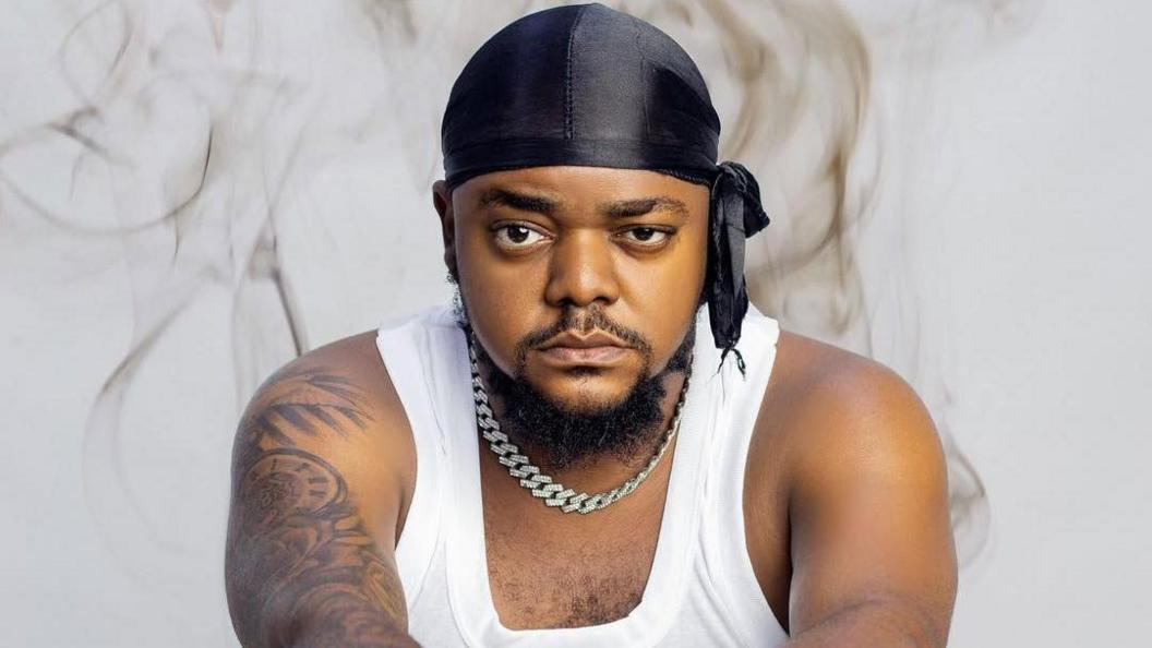 Odira Nwobu poses in a white vest and black durag. His expression is serious and thoughtful.