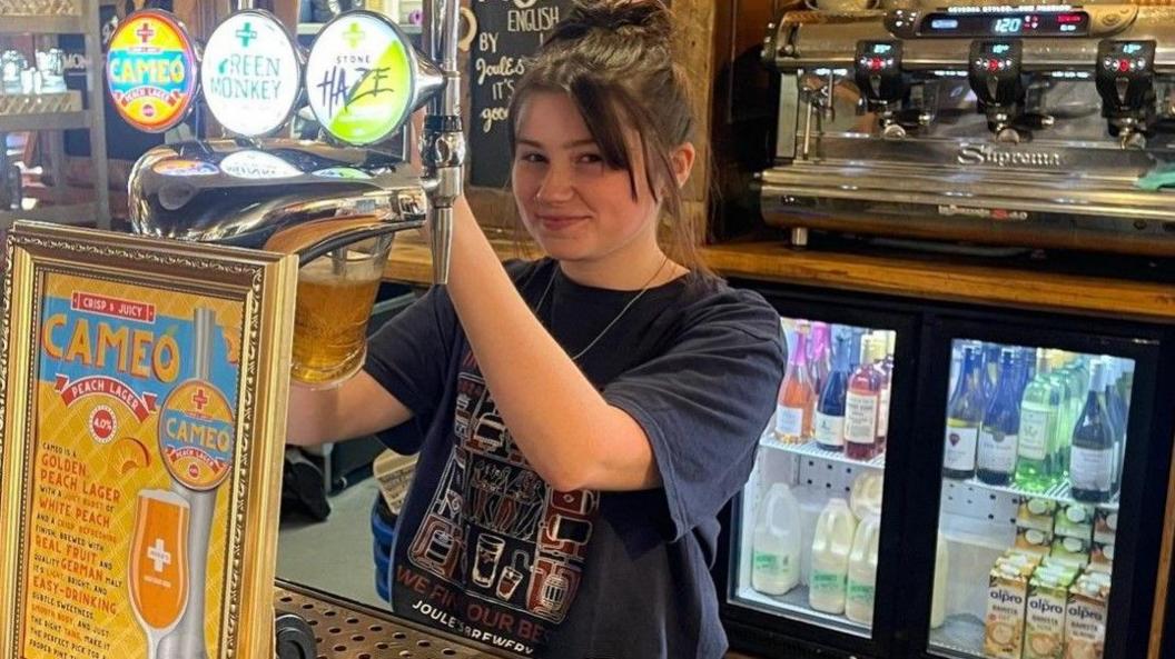 A barmaid is pulling a pint at the bar. Drinks are in a fridge behind her.