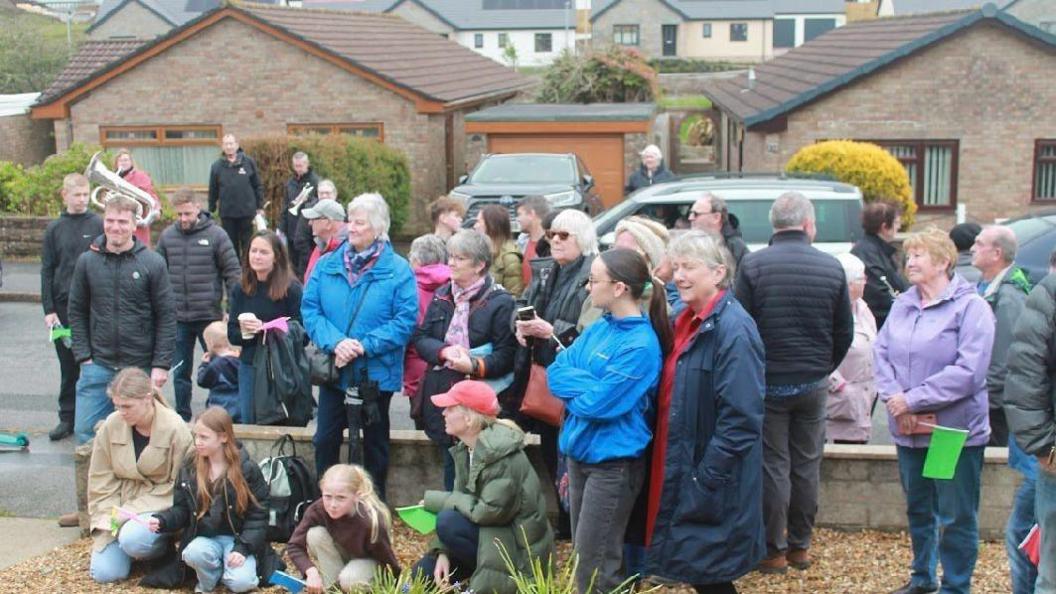 Dozens of people stand outside Nancy Tresidder's house for her 100th birthday party. Most are wearing coats and some are carrying green or blue flags. Members of a brass band are in the background.