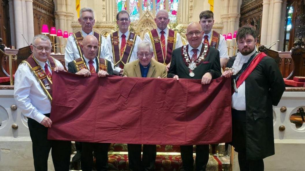 A group of men stand at the front of a church. Most of them are dressed in white shirts and ties, and are wearing the crimson sash of the Apprentice Boys of Derry. Four of the men are at the back of the image, with a further five in front of them - the men in front are holding onto a crimson flag which is being presented to a clergy man.