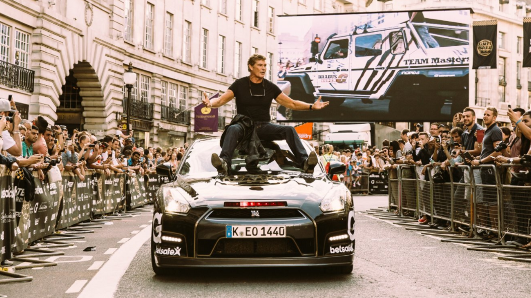 A man in all black clothing sits in on top of a black sports car with crowds either side of him.