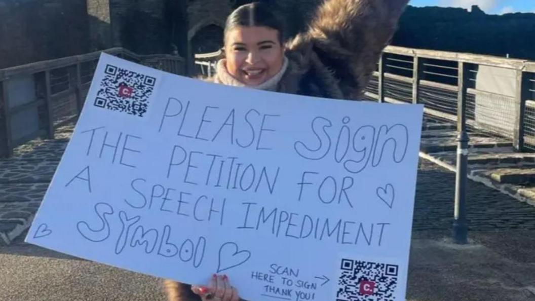Jessie Yendle holding white placard with QR codes and message saying please sign the petition for a speech impediment symbol