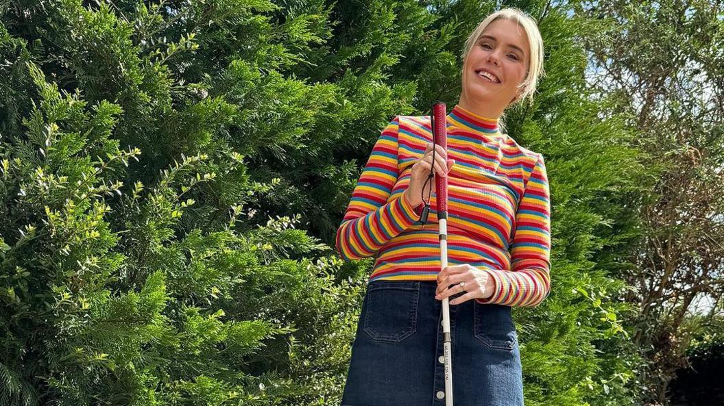 Claire Sisk wearing a bright, multi coloured top, with a blue denim skirt. She is smiling to the camera and she is slightly leaning to the right. She has her hands on a white cane and has a ring on her finger. Her blonde hair is tied back. She is standing in front of a green foliage and is outside.